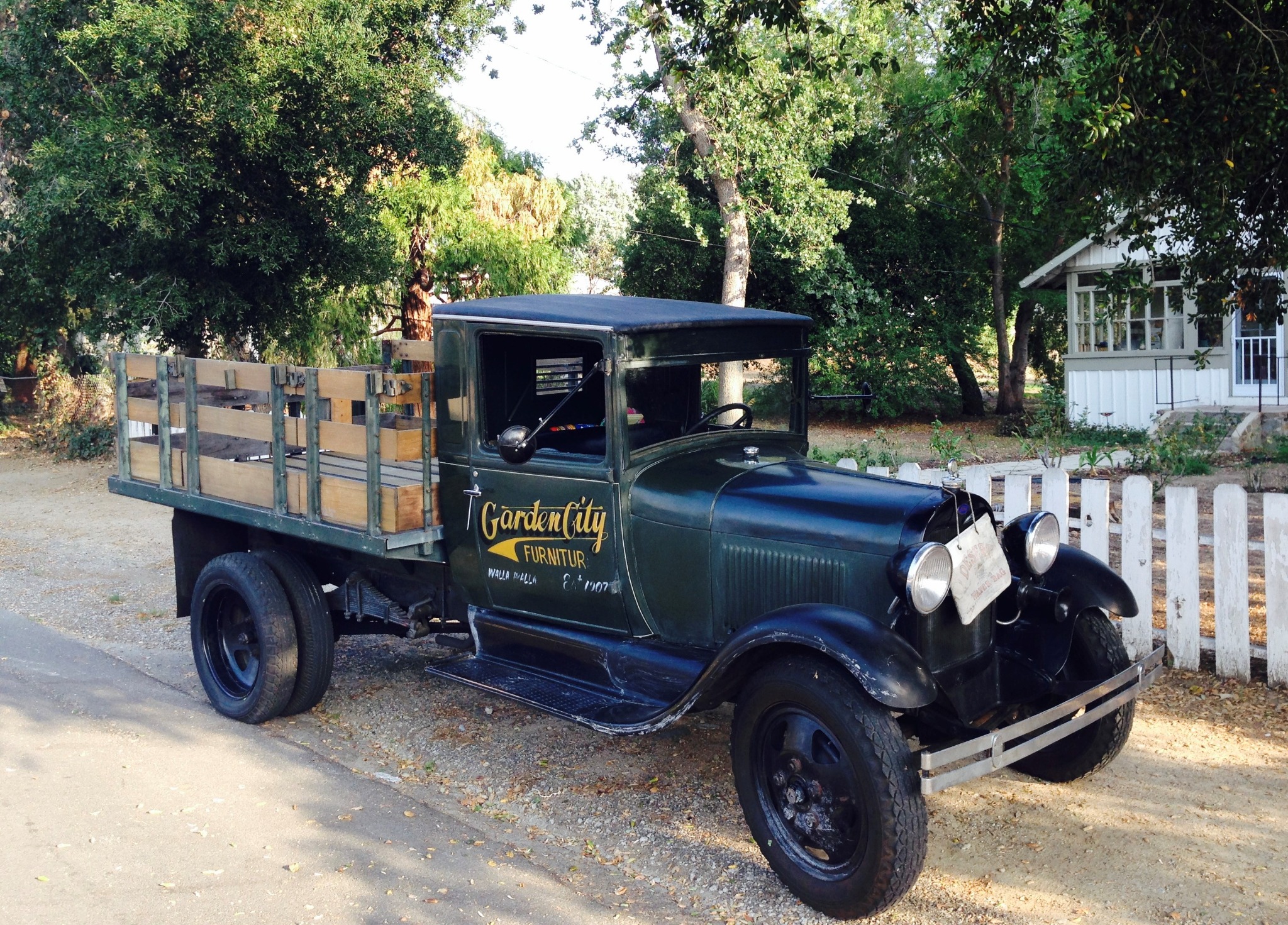 Green Ford Model AA stake-bed truck with Garden City Furniture lettering parked beside a white fence.